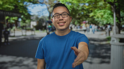 Young chinese man in a blue t shirt offers his hand for a handshake on a busy city street while...