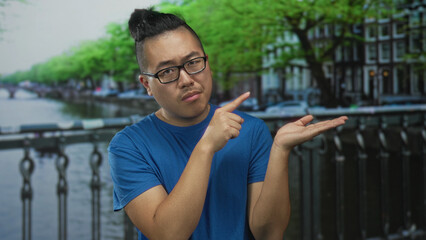 Man pointing finger to open palm on street beside amsterdam canal bridge, wearing blue tshirt and...