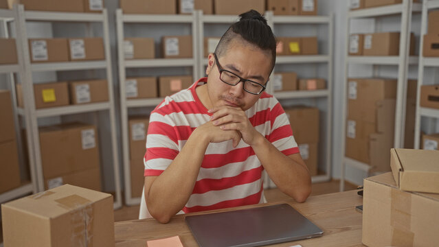 Young chinese man covering mouth with hand while seated at laptop among stacked parcel boxes inside a packing building; concern.