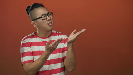 Young man with open palms gesture and glasses in studio against an orange wall wearing a red and white striped tee, looking puzzled; confusion.