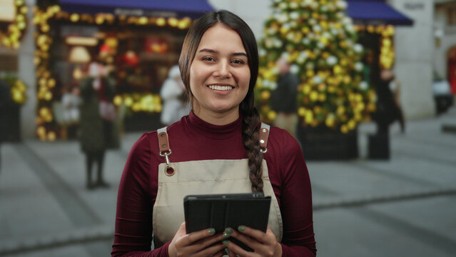 Woman smiling in apron holding tablet in festive decorated outdoor street with blurred christmas tree and storefronts, showcasing holiday spirit and outdoor interaction. - Powered by Adobe
