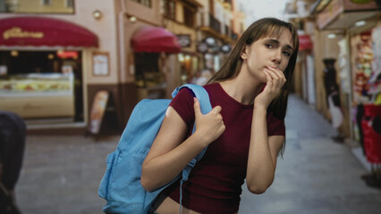 Young woman holding backpack strap on a narrow city street, hand to mouth thoughtful gesture;...