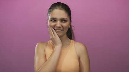Teen girl wearing orange tank top holds cheek with hand and winces in pink studio; toothache severe pain.
