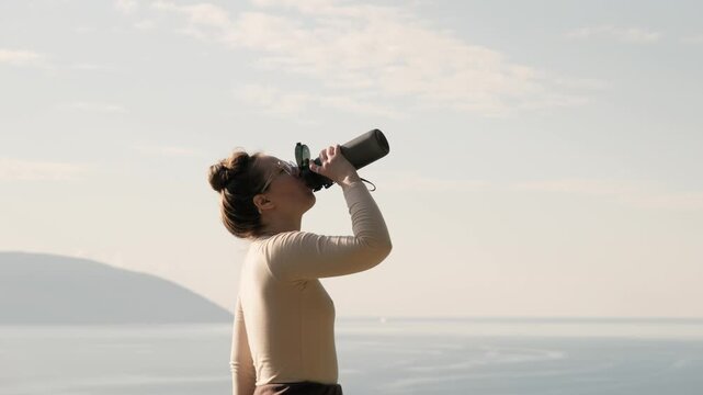 Woman drinking water outdoors by the sea, quenching thirst after fitness or yoga practice. Healthy lifestyle, hydration, wellness and recovery in calm natural coastal setting.