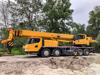 Yellow construction crane on a dirt area with sparse vegetation, overcast sky, trees in the distance, soft shadows, lattice boom structure, multiple axles, enclosed cab, heavy machinery © Mister