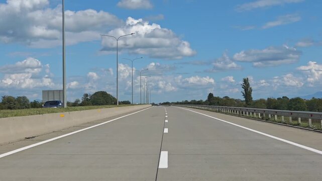 The perspective is from inside a vehicle cruising along a wide multi lane freeway. The sun is shining brightly in a clear blue sky with fluffy white clouds scattered above the highway