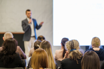 Presenter speaks to audience in conference room with bright screen behind. Audience seated listening attentively.