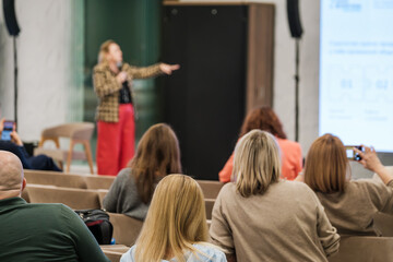 A speaker presents to an audience in a conference room with a projector screen. Attentive attendees listen and some capture the moment with phones.