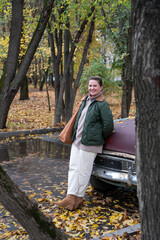Fototapeta premium Smiling woman stands by a vintage turquoise car in a quiet autumn courtyard, fallen leaves, muted green and yellow tones, calm urban atmosphere