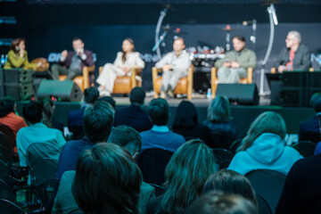 Panel discussion on stage with diverse speakers and attentive audience. Conference atmosphere with live talk and engaged crowd. Audience sits in rows watching panel lights highlight participants 