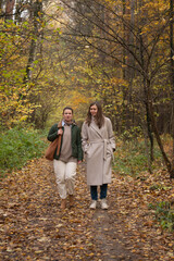 Fototapeta premium Two women walk together along a forest path covered with autumn leaves, tall trees around, natural daylight, relaxed and friendly atmosphere