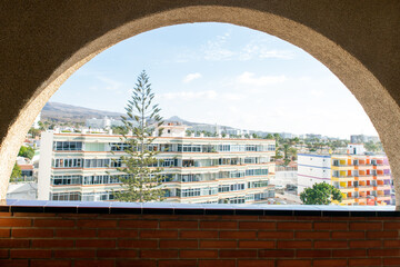 Photograph taken through shaped window, semi-circular one, which frames a view of modern high-rise buildings. A red brick wall section Outside features multi-story buildings with numerous windows. 