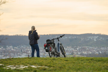 Obraz premium Cyclist on hilltop overlooking city. Person with bicycle on hilltop with gear, overlooking city, forest and evening sky.