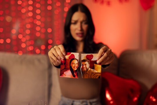 Unhappy young woman ripping photo with ex-husband and crying, sitting on sofa at home on Valentine's day