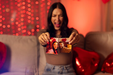 Furious woman ripping photo with ex-husband and shouting, sitting on sofa at home on Valentine's day