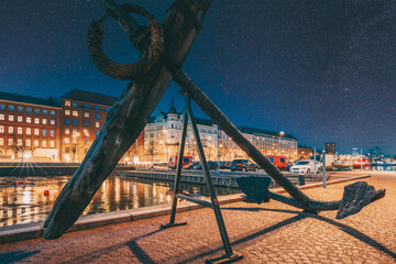 Helsinki, Finland. Old Anchor On Pier With View Of Pohjoisranta Street In Evening Night Illuminations. Night stars shining above street