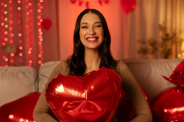 Happy woman celebrating Valentine's day, sitting on sofa holding large red heart-shaped balloon, smiling broadly, showing happiness in festive setting