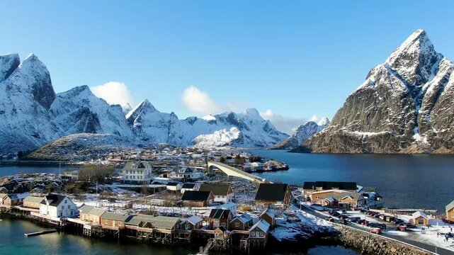A drone soars above the snow-covered Lofoten landscape at Sakris&oslash;y near Reine, where sunlit rorbuer cabins contrast beautifully with mountains and icy waters.