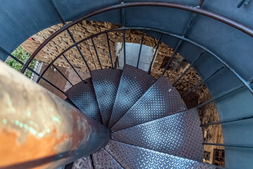 Overhead view of a modern spiral staircase with metal diamond-plate steps and black railings inside a historic stone building, highlighting geometric patterns against rustic ancient walls