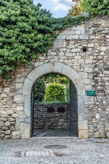 Fototapeta premium Open iron gate to Jardins de la Francesa in Girona through an arched stone doorway covered in ivy. Cobblestone path leads into lush garden beyond medieval walls. Girona, Spain, Nov 28, 2021
