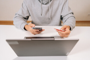 Cropped shot of person making online purchase, holding smartphone and red credit card, using laptop to complete secure e-transaction, focused on digital payment for convenient shopping.