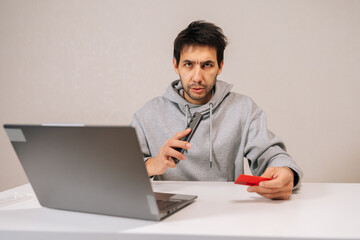 Portrait of bearded young man in hoodie making online purchase, holding phone and credit card, using laptop to complete secure e-transaction, focused on digital payment for convenient shopping.