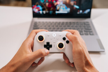 Person holding white wireless game controller with illuminated joysticks and buttons. Interacting with laptop in background. Symbolizing online gaming. Connectivity and digital entertainment.