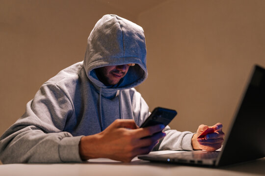 Hooded person engaging in cybercrime, performing online fraud and data theft using mobile phone, laptop and credit card in dark environment, representing internet security threats. - Powered by Adobe