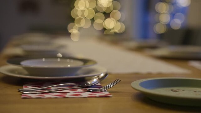A festive Christmas table setting features white plates with textured blue rims on a wooden table. A holiday-themed runner, napkins with wreaths, and silver cutlery create a warm, inviting scene.