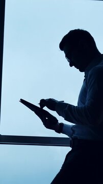 Side view of a freelancer sitting on the desk. Man in glasses uses stylus pencil working on I-pad. Low angle view. Wide window at backdrop. Vertical video