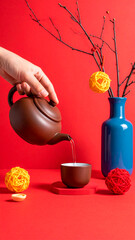 A hand pouring tea from a traditional brown teapot into a small cup. A tall blue vase with bare branches holds red and yellow woven ornaments, with more on the surface. Ideal for Lunar New Year themes