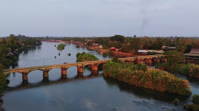 Aerial view of the historic French colonial bridge connecting Don Det and Don Khon islands over a tranquil river, surrounded by lush tropical vegetation and traditional houses in Laos.