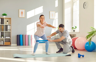 Positive woman in a rehabilitation fitness session with personal trainer in gym. Focused on squats exercise with a band, the male coach guides her workout. 
