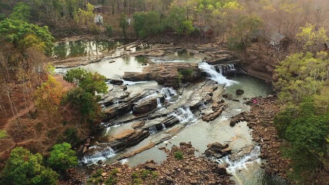 Scenic aerial shot of Tad Lo waterfall cascading over rocky terrain surrounded by lush and dry forest in southern Laos.