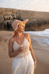 Young woman in white summer dress wearing hat holding wineglass on sandy beach near sea enjoying vacation with rocky coastline during sunny day