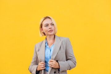 Confident blond businesswoman in stylish blazer standing against bright yellow background looking thoughtful with modern professional appearance