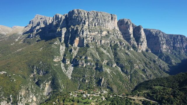 Aerial view of dramatic cliffs and a small village at Vikos Gorge in Greece, under a clear blue sky. Lush green forests cover the landscape, highlighting the natural beauty of the region.