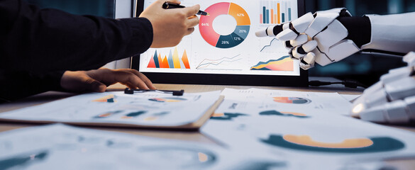 A human hand collaborates with a robotic hand to analyze data charts displayed on a screen, highlighting the integration of technology in modern business analytics.Noogenesis