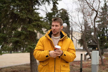 Fototapeta premium Man stands in park wearing yellow jacket and holds a coffee cup in his hands