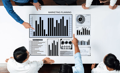 A business team engages in collaborative marketing planning, analyzing charts and graphs on a large table. This scene captures teamwork and strategic discussions in a modern office environment. Axiom