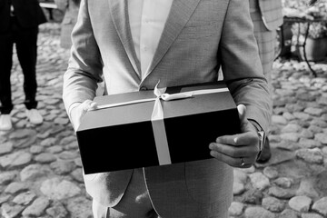 Black-and-white photo of a well-dressed man holding an elegant gift box tied with a ribbon, symbolizing celebration, generosity, and formal occasion moments.