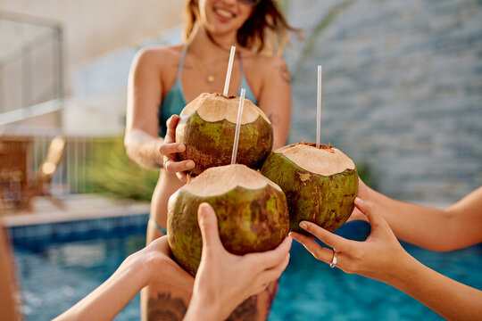 Group of friends raising tropical coconut drinks with straws while relaxing by the pool on a summer vacation day
