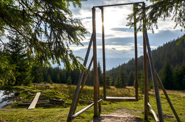 Rustic wooden swing on a grassy mountain clearing, framed by dark trees. Sun flares through the clouds and the wooden structure, overlooking a dense evergreen forest. Carpathians