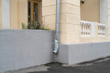 Water drain pipe runs down the corner of a building with some plants nearby on a gray wall