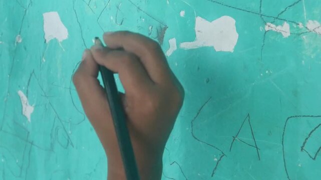 A child's hand holding a pencil and drawing creative scribbles on a green wall surface.
