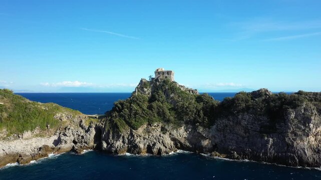 Dramatic rocky coastline with lush greenery and a historic stone tower overlooking the blue sea at Capo di Conca, Italy. Clear sky and vibrant Mediterranean landscape create a scenic view.