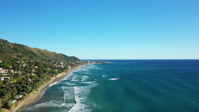 Scenic aerial view of the Acciaroli coastline with turquoise sea, sandy beach, and lush green hills under a clear blue sky in southern Italy.