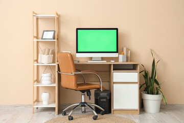 Modern computer with blank screen on table and stylish armchair near beige wall in home office