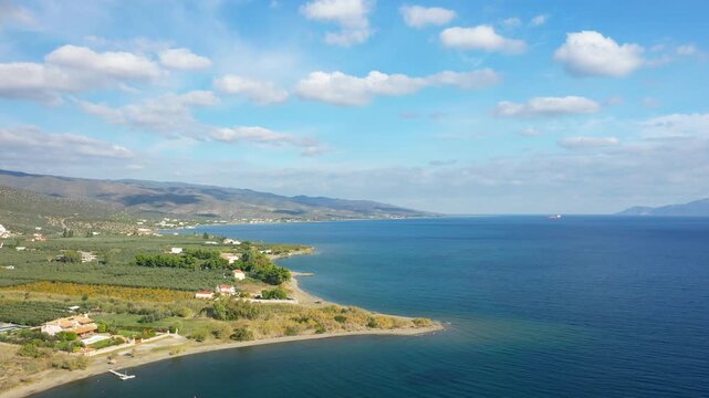 Aerial view of a serene coastline with clear blue water, green fields, and scattered houses near Hermione, Greece under a partly cloudy sky.