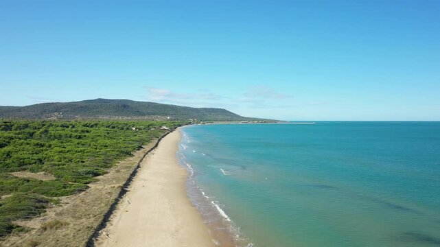 Drone shot of a pristine sandy beach and turquoise sea along the Isola Varano natural reserve, with lush greenery and distant hills under a clear blue sky.